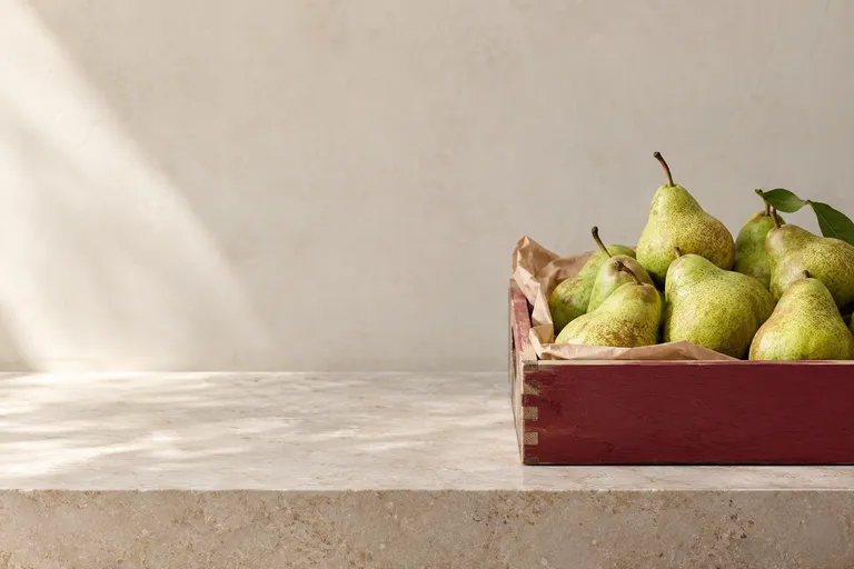 A beautifully ordered grocery produce display with a few fresh green pears and one burgundy crate edge, abundant pale counter and empty wall, no pricing or signs.