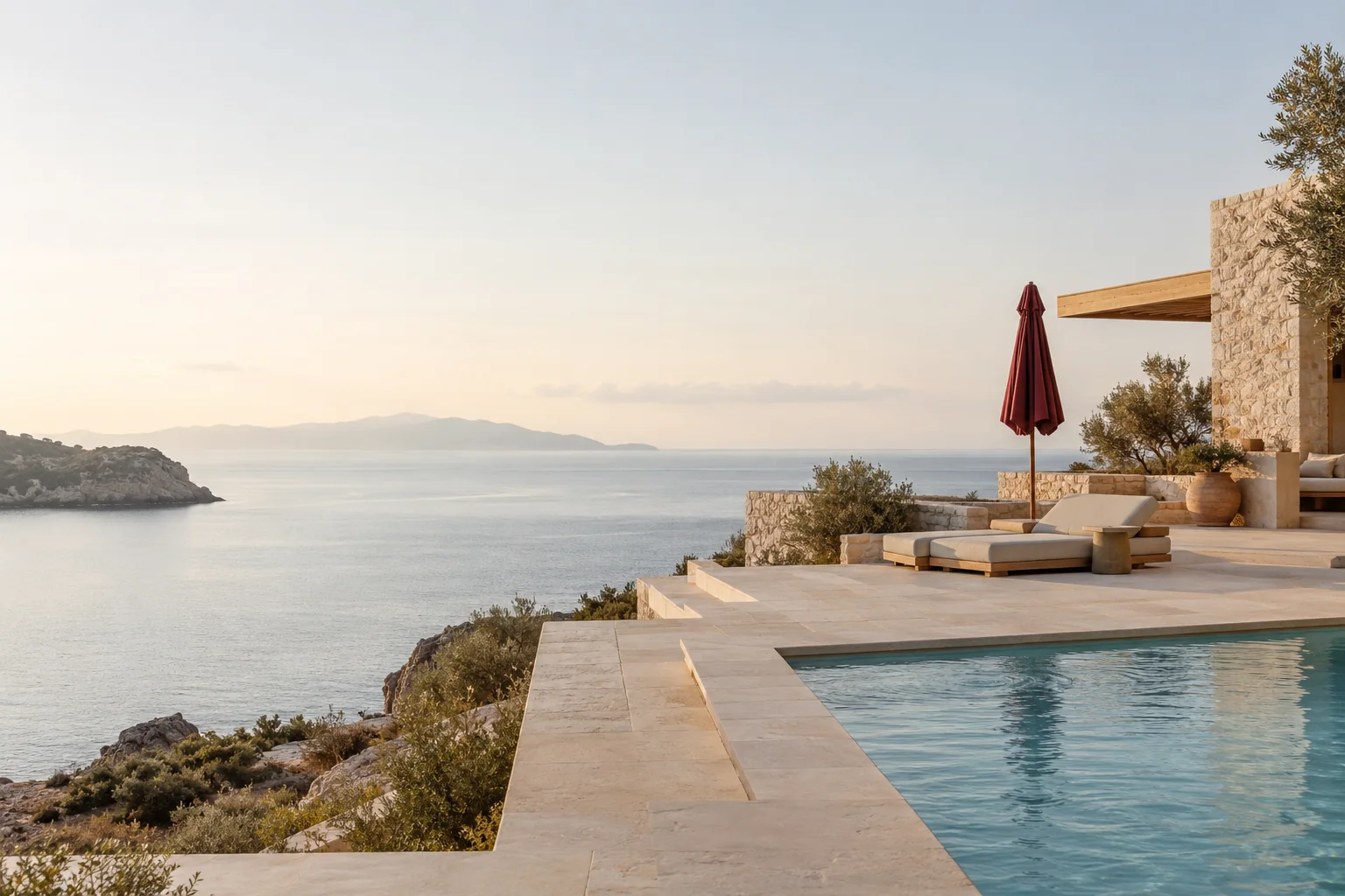 An understated destination resort on a quiet coastline, pale stone terraces, low rise architecture and a serene pool at the right, a single closed burgundy parasol, broad empty pale sky and calm sea, no people.