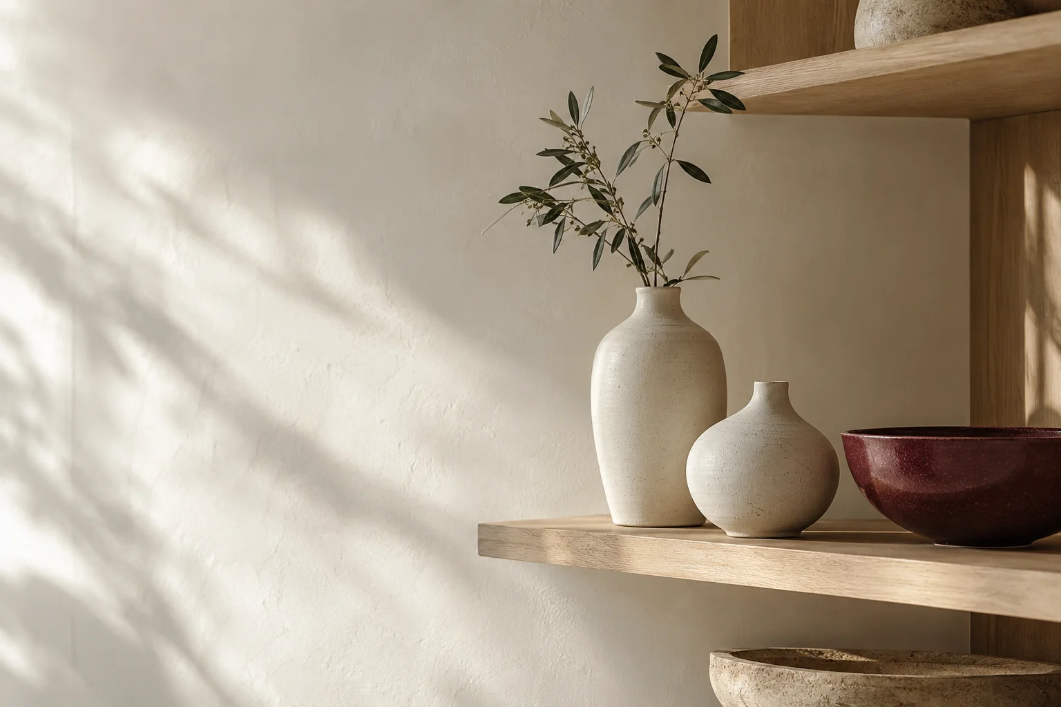 A restrained homeware specialty shop display with two ivory ceramic vessels and a burgundy ceramic bowl on pale oak shelving, generous uncluttered wall space.