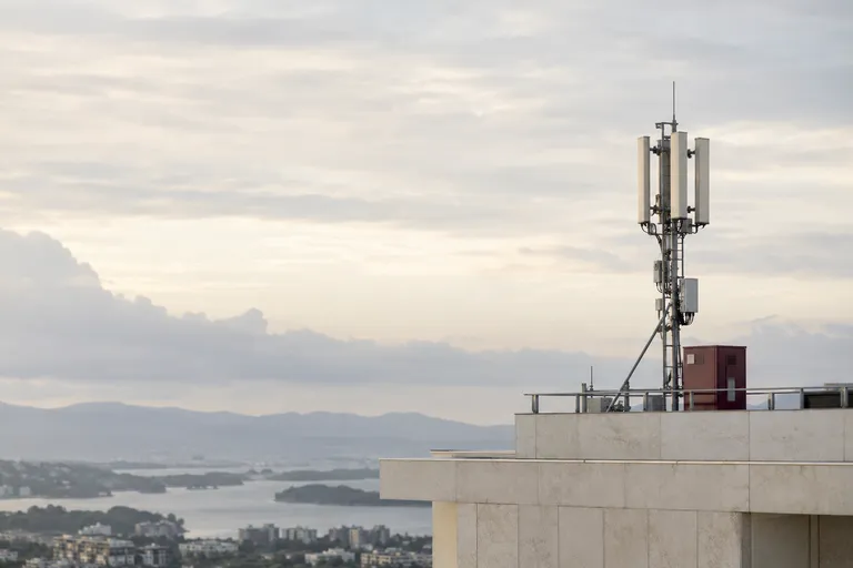 A slender real telecom antenna installation on a distant pale rooftop with a small burgundy maintenance cabinet, expansive soft overcast sky and quiet geometry.