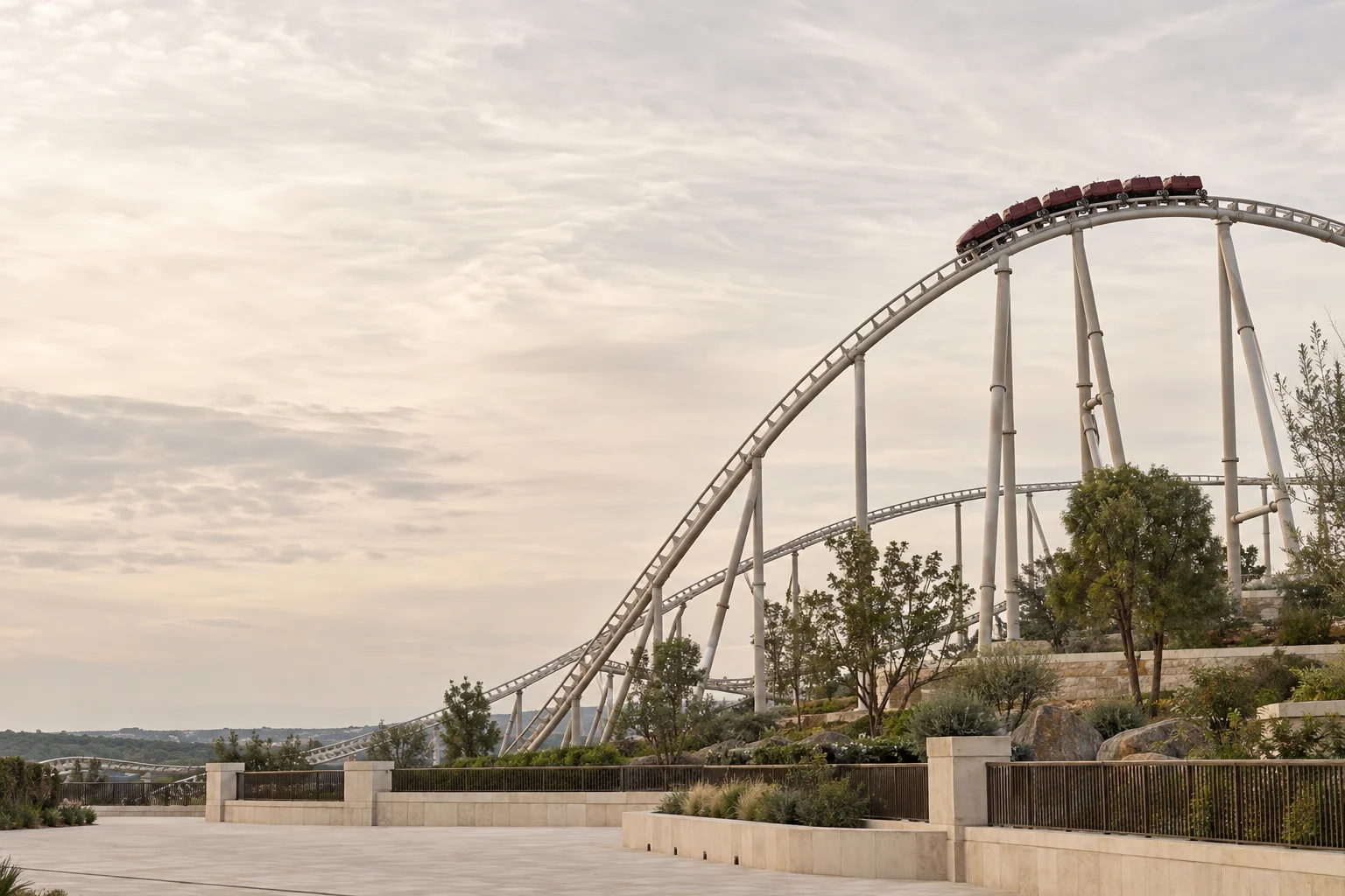 A pale steel roller coaster with a burgundy train above a quiet landscaped park, framed by an expansive soft sky.